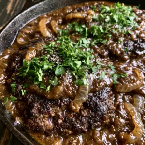 Extreme close-up of seared hamburger steaks with dark crust and jammy caramelized onions in a black skillet.